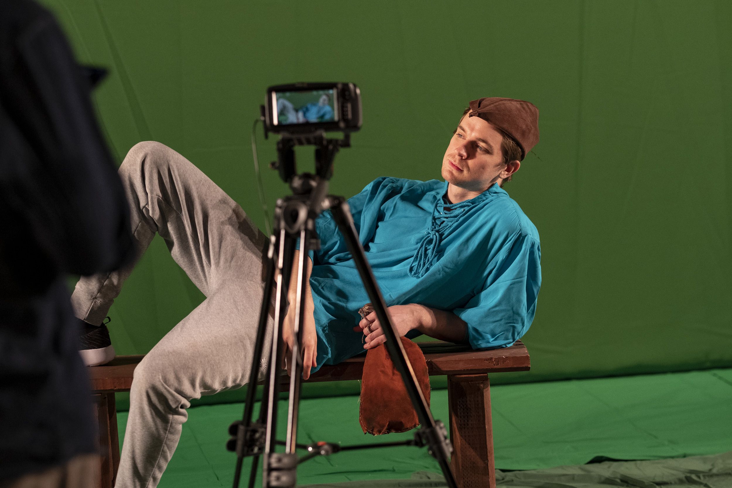 An actor in a blue shirt and brown hat lays on a bench, posing for the camera..