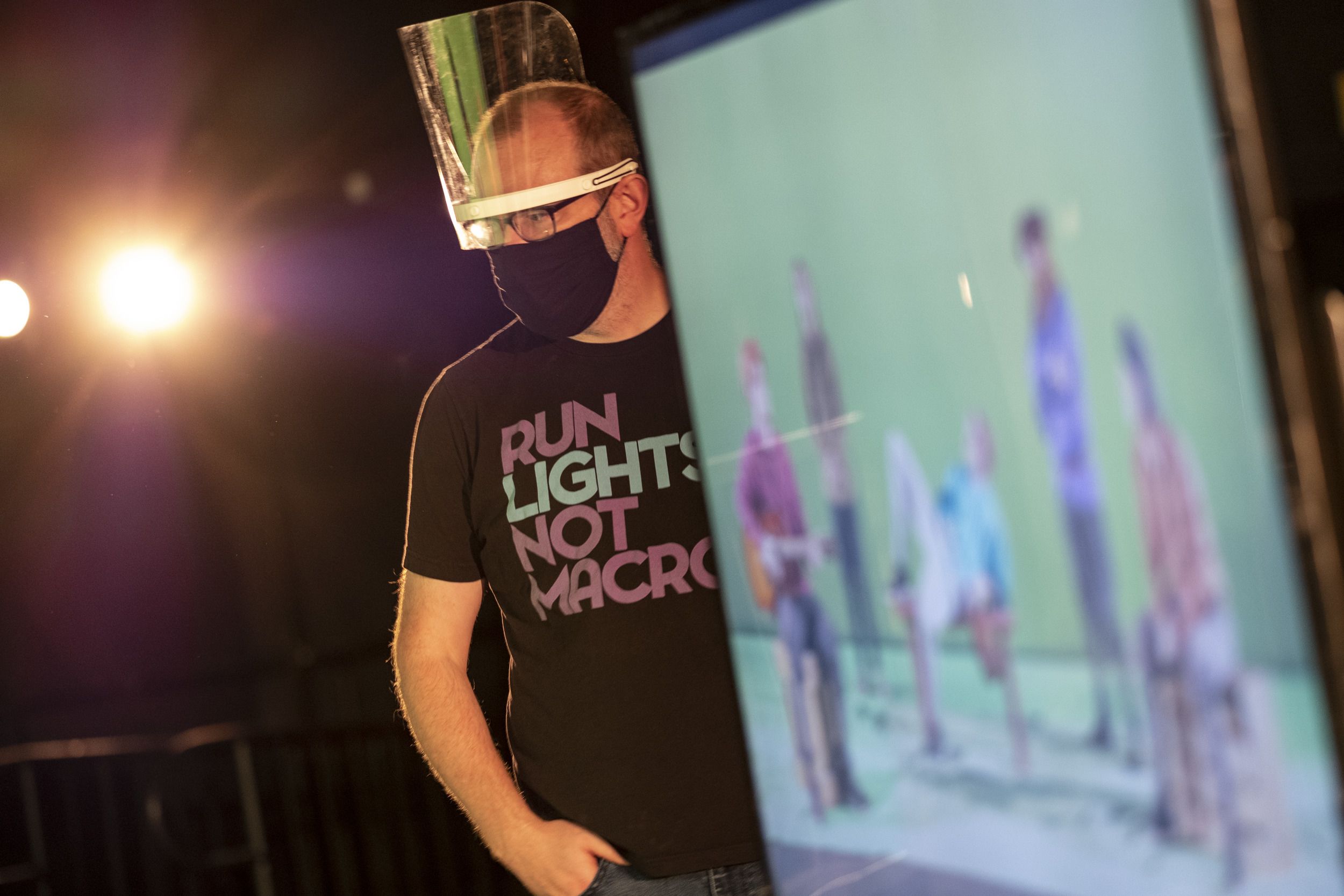 A screen displays the actors on set. Beside the screen is a crew member with a face shield.